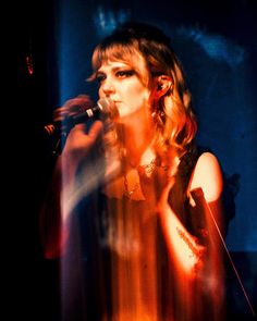 a woman singing into a microphone in front of a dark background with light coming from behind her