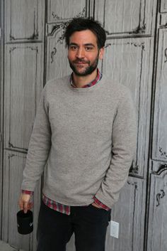 a man standing in front of a wall wearing a gray sweater and black pants with his hands in his pockets