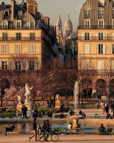 people are walking around in the park with many buildings behind them and a fountain surrounded by trees