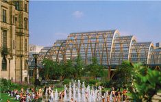 a group of people standing around a fountain in the middle of a park with lots of glass buildings behind it