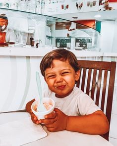 a little boy sitting at a table eating something out of a paper cup in front of him