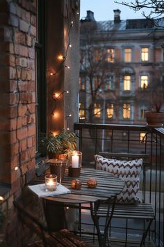 Cozy balcony with string lights, candles, and a pillow on a chair, overlooking city buildings at dusk. Witchy Balcony, Balcony Makeover Ideas, Vintage Balcony, Balcony Makeover