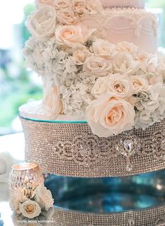 a three tiered wedding cake with white flowers on the top and side, surrounded by crystals