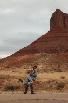 two people walking down the road in front of a mountain with a tall rock formation behind them