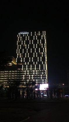 people standing in front of a tall building with lights on it's side at night