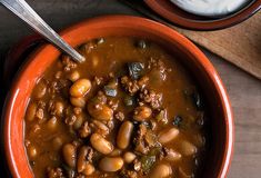 a bowl filled with beans and meat on top of a wooden table next to a spoon