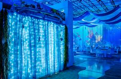 a room filled with lots of white tables and chairs covered in blue lights next to tall drapes