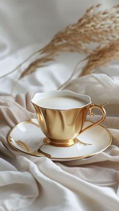 a golden coffee cup and saucer on a white bed sheet with dried grass in the background