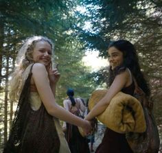 two young women dressed in medieval clothing holding hands and smiling at each other while walking through the woods