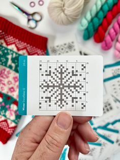 a person holding up a piece of paper with a crossword pattern on it next to some knitting supplies