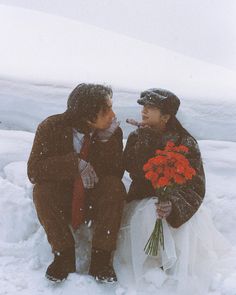 two people are sitting in the snow and one is holding a bouquet of red flowers