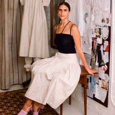 a woman sitting on top of a wooden chair in front of a dress hanging up