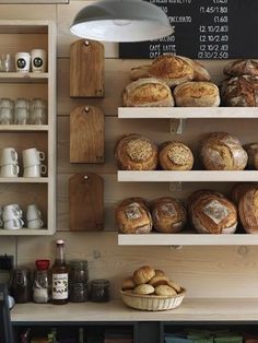 breads and rolls are on shelves in a bakery with a chalkboard behind them