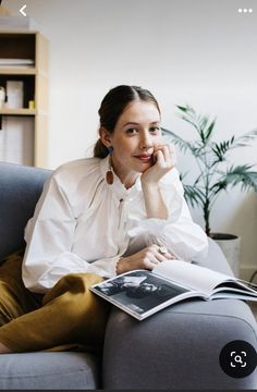 a woman sitting on a couch reading a book and looking at the camera with her hand under her chin