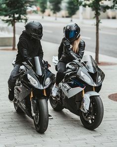 two people riding on the back of motorcycles down a brick road with trees in the background