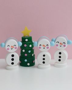 three snowmen next to a christmas tree on a white table with pink wall in the background