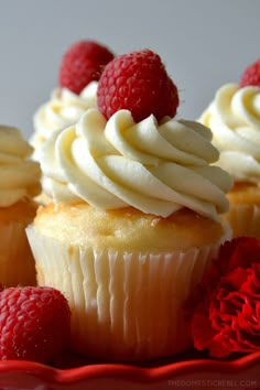 three cupcakes with white frosting and raspberries on top, sitting on a red plate