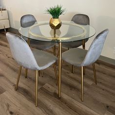 a glass dining table with four chairs and a gold vase on the top, in front of a white wall