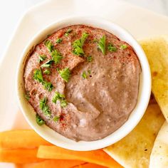 a white plate topped with a bowl of dip next to sliced carrots and pita chips