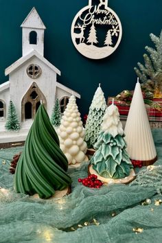 three small christmas trees sitting on top of a table next to a white church and blue wall