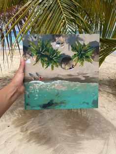 a person holding up a painting on the beach with palm trees in the foreground