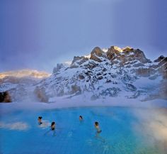 three people are swimming in a blue pool with mountains in the background and snow on the ground