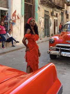 a woman in a red dress standing next to an orange car on a street with people sitting around