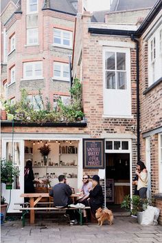 people are sitting at tables outside in front of a building with plants growing on the roof