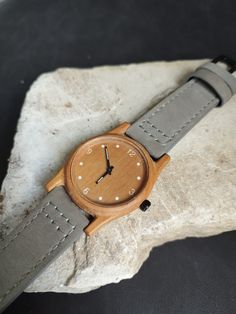 Experience the fusion of tradition, craftsmanship, and style with this elegant wooden wristwatch, handcrafted from premium teak wood. Our watch is a testament to the timeless allure of natural materials, blending together metal, wood, and leather to create an accessory that is as stylish as it is sustainable. The watch's body is fashioned from teak wood, a material beloved for its rich color, durability, and unique grain patterns. Each watch is a work of art with its own unique wood grain, ensuring that no two pieces are exactly alike. The classic style of the watch is accentuated by white numbers displayed on a clear, easily readable watch face. A soft, gray leather strap adds a touch of sophistication and ensures a comfortable fit. The strap is adjustable and sturdy, ideal for everyday w Gray Leather, Wristwatch Men, Unique Charms, Teak Wood, Grey Leather, Accessories Unique, Wood Watch, Leather Material, Natural Materials