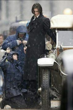 a man in a trench coat standing next to an old car with another man sitting on it