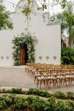 rows of wooden chairs sitting in front of a white building with greenery around it