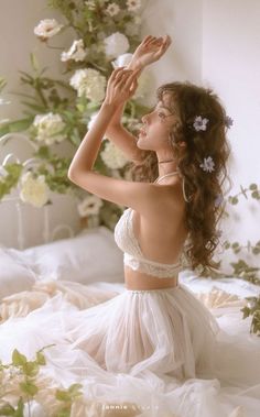 a woman in a white dress is sitting on a bed with flowers and greenery