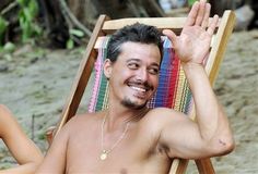 a shirtless man sitting in a chair on the beach with his hand up to his head