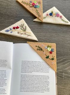 an open book sitting on top of a wooden table next to three pieces of embroidered fabric