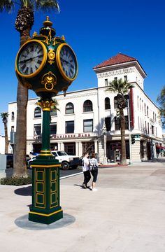 there is a clock on the pole in the middle of the street with people walking by