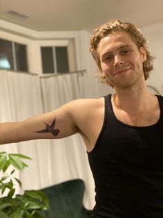 a man with a bird tattoo on his arm in front of a green couch and potted plant