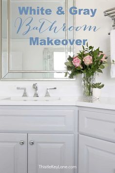 a white bathroom with blue and gray lettering on the mirror over the sink is a vase full of flowers