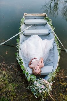 a woman in a white dress is laying on a boat with greenery around her