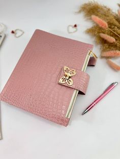 a pink notebook with a gold bee on the cover and some pens next to it