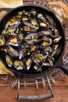 a skillet filled with mussels on top of a wooden table next to bread