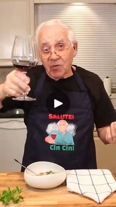 an old man holding a glass of wine in his hand while standing at a kitchen counter