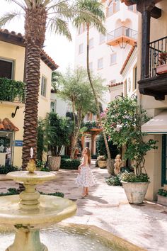 a woman standing in front of a fountain surrounded by palm trees and potted plants