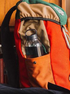 an orange and green backpack with a water bottle in it's pocket next to a book shelf