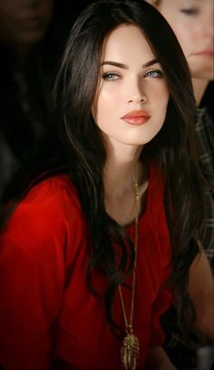 a woman with long dark hair wearing a red shirt and gold necklace sitting in front of other people