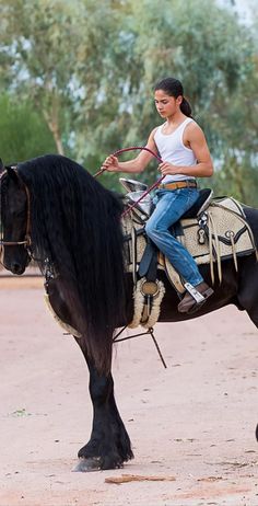 a woman riding on the back of a black horse in a dirt field next to trees