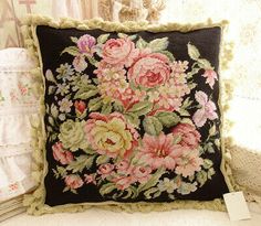 a black pillow with pink and yellow flowers on it sitting next to a white lace curtain