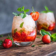 two glasses filled with ice and strawberries on top of a wooden table next to mint leaves