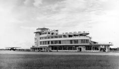 black and white photograph of an airplane parked in front of a building with multiple stories