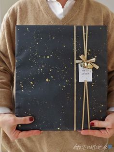 a person holding a gift wrapped in black paper and tied with twine, while wearing a brown sweater