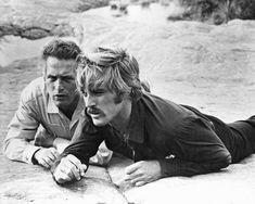 two men laying on the ground in front of some rocks and one is looking at something